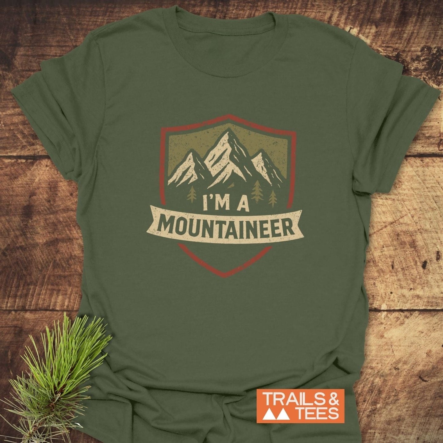 Mountaineer T-Shirt with badge graphic of peaks and trees on olive-green fabric, showcasing “I’m a Mountaineer” banner, emphasizing outdoor adventure.