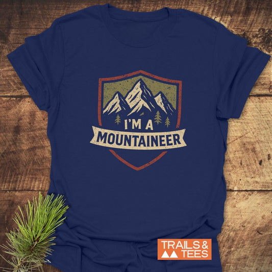 Mountaineer T-Shirt showcasing a mountain-and-trees graphic inside a shield with a banner on navy-blue fabric, laid flat beside an orange TRAILS & TEES logo.