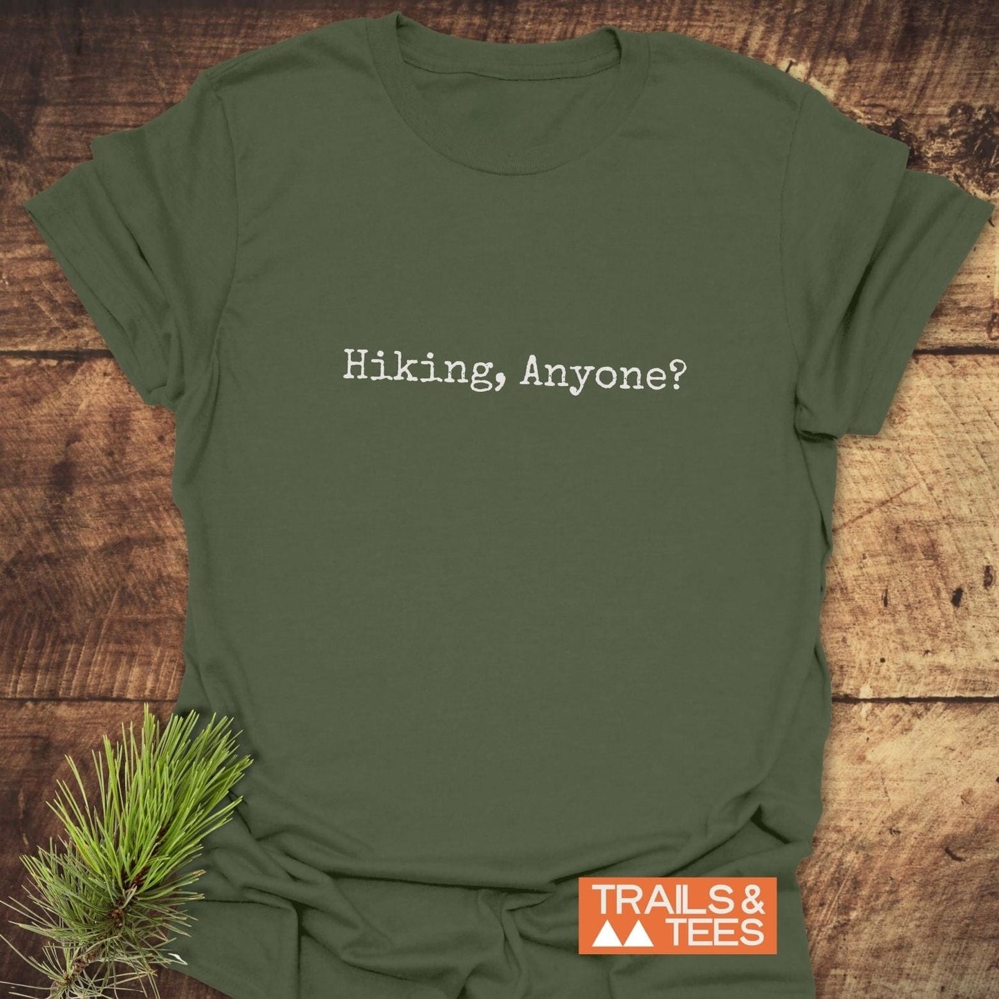 Olive-green Hiking, Anyone? T-Shirt in typewriter font on wood, orange TRAILS & TEES logo, pine sprig; crafted from soft, durable ring-spun cotton.