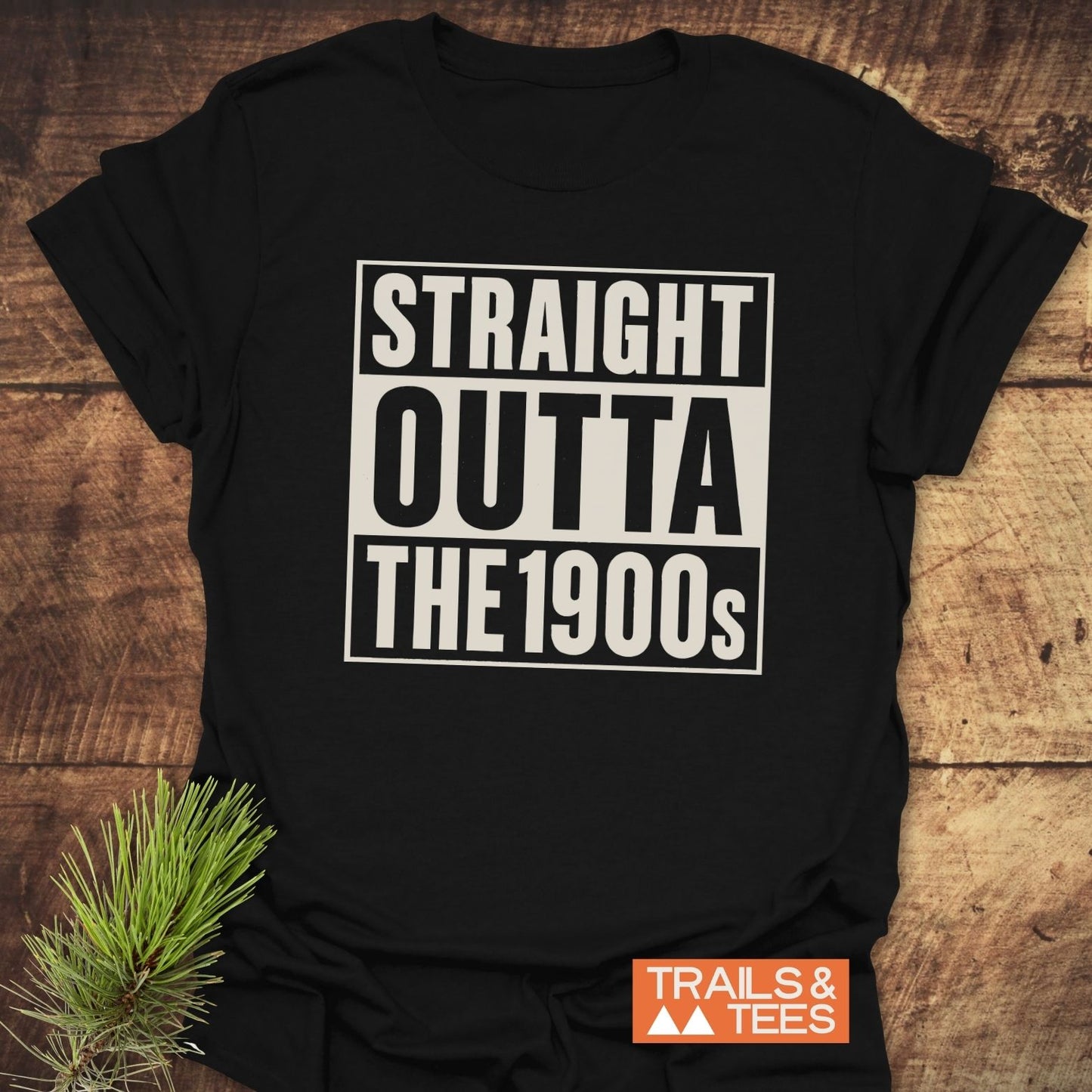 Straight Outta The 1900s T-Shirt on wooden surface with pine sprig, featuring bold white text. Made from soft, ring-spun cotton for comfort and durability.