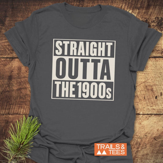 Charcoal-gray T-shirt with STRAIGHT OUTTA THE 1900s graphic, featuring a small TRAILS & TEES logo. Laid on wooden surface with a pine sprig accent.