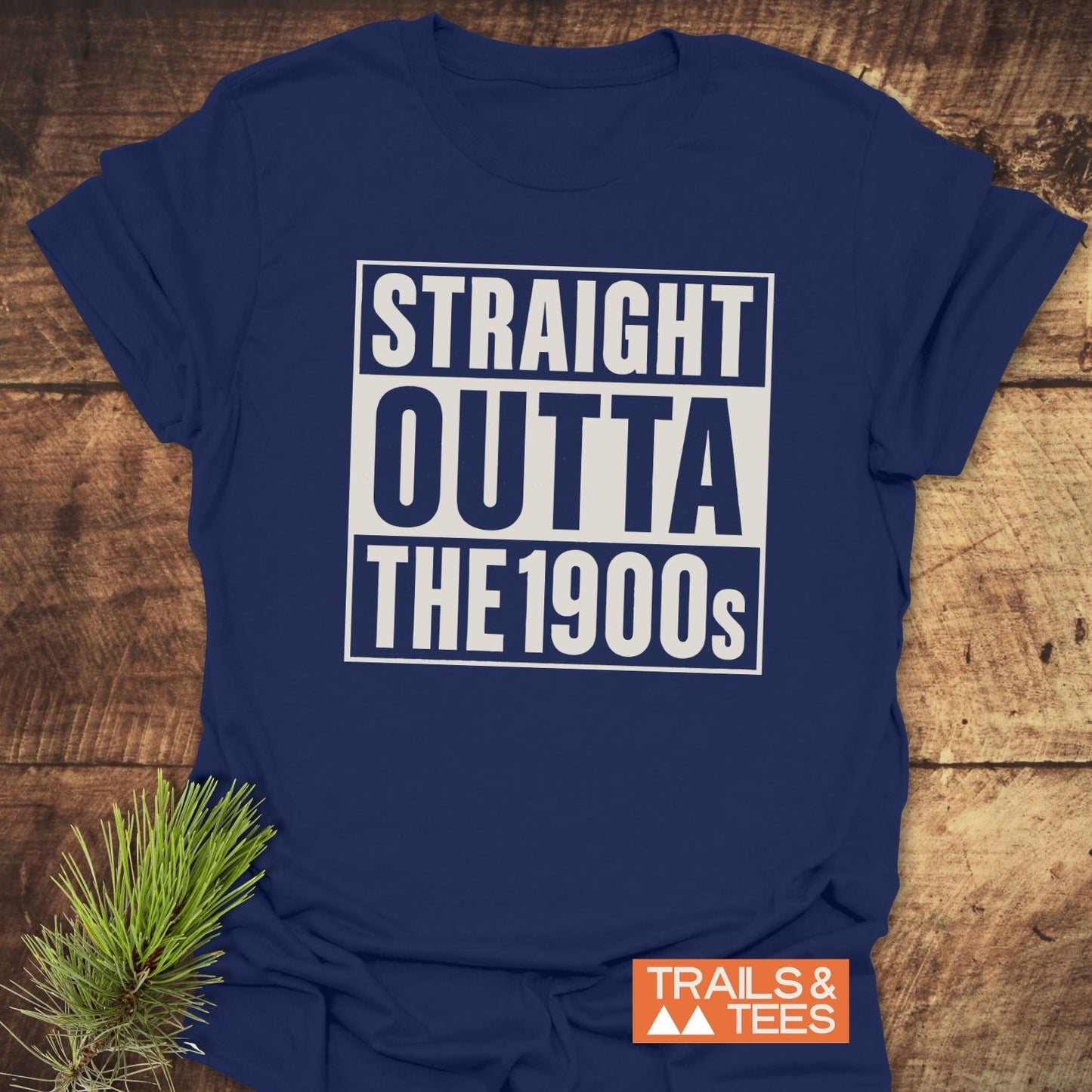 Navy-blue Straight Outta The 1900s T-shirt on wood, featuring durable white print, Trails & Tees logo, styled with a pine branch for outdoor appeal.