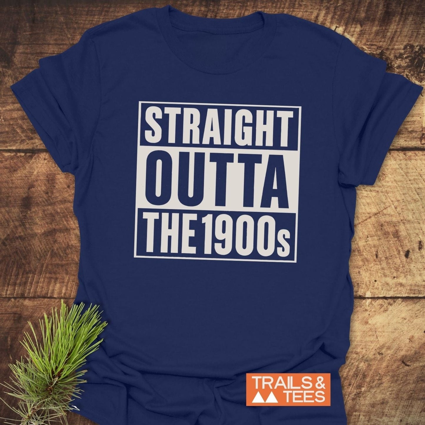 Navy-blue Straight Outta The 1900s T-shirt on wood, featuring durable white print, Trails & Tees logo, styled with a pine branch for outdoor appeal.