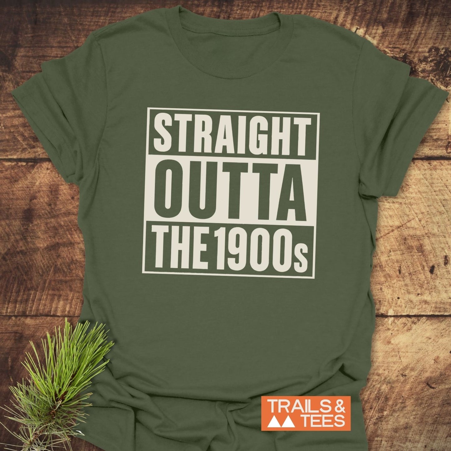 Straight Outta The 1900s T-Shirt in olive-green, displayed flat with bold text, rustic background, pine sprig, and Trails & Tees logo.