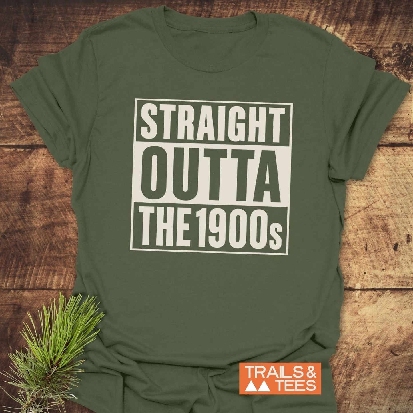 Straight Outta The 1900s T-Shirt in olive-green, displayed flat with bold text, rustic background, pine sprig, and Trails & Tees logo.