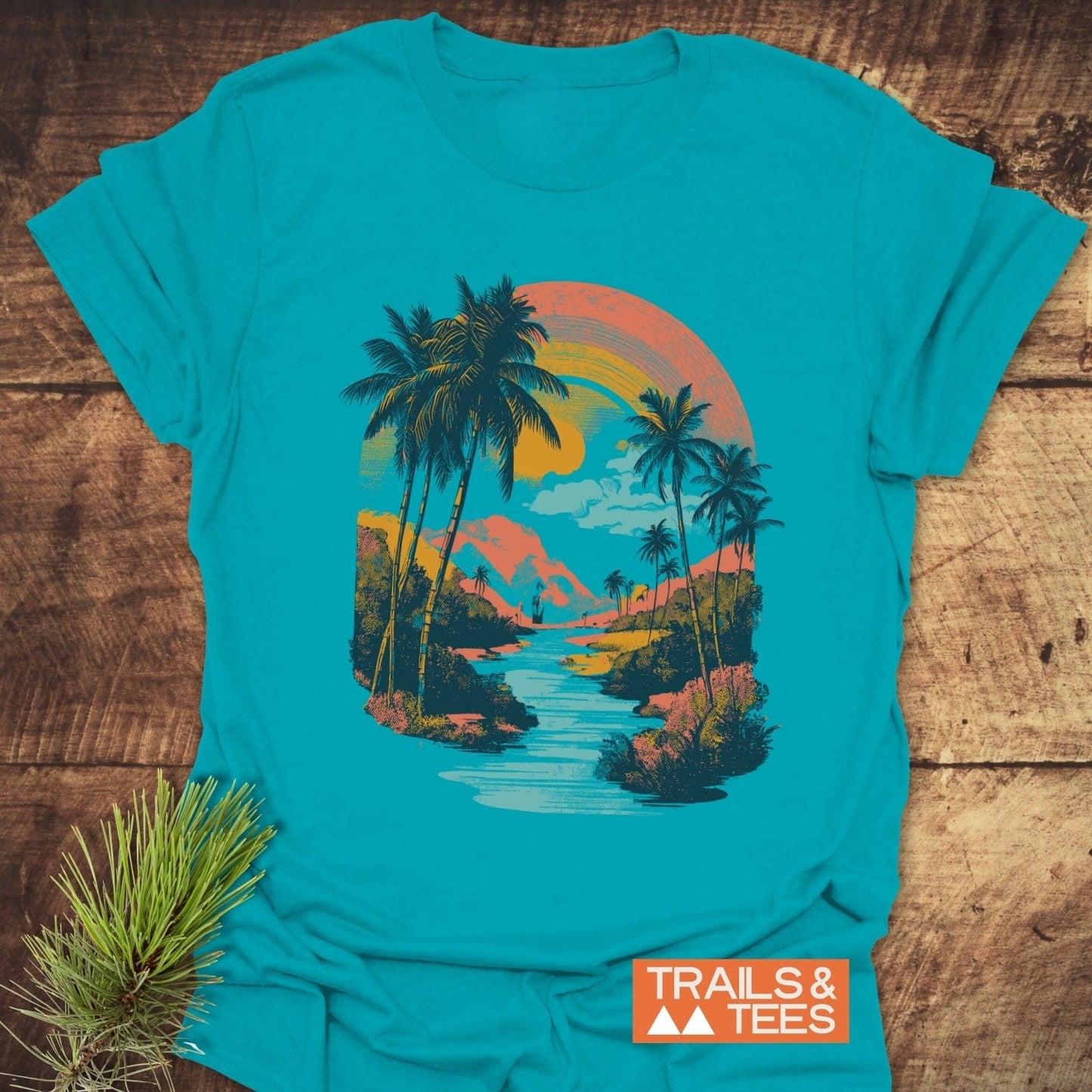 Along The River T-Shirt featuring a tropical landscape with palm trees and sunset hues, crafted from soft cotton for comfort and durability, logo “TRAILS & TEES.” visible.