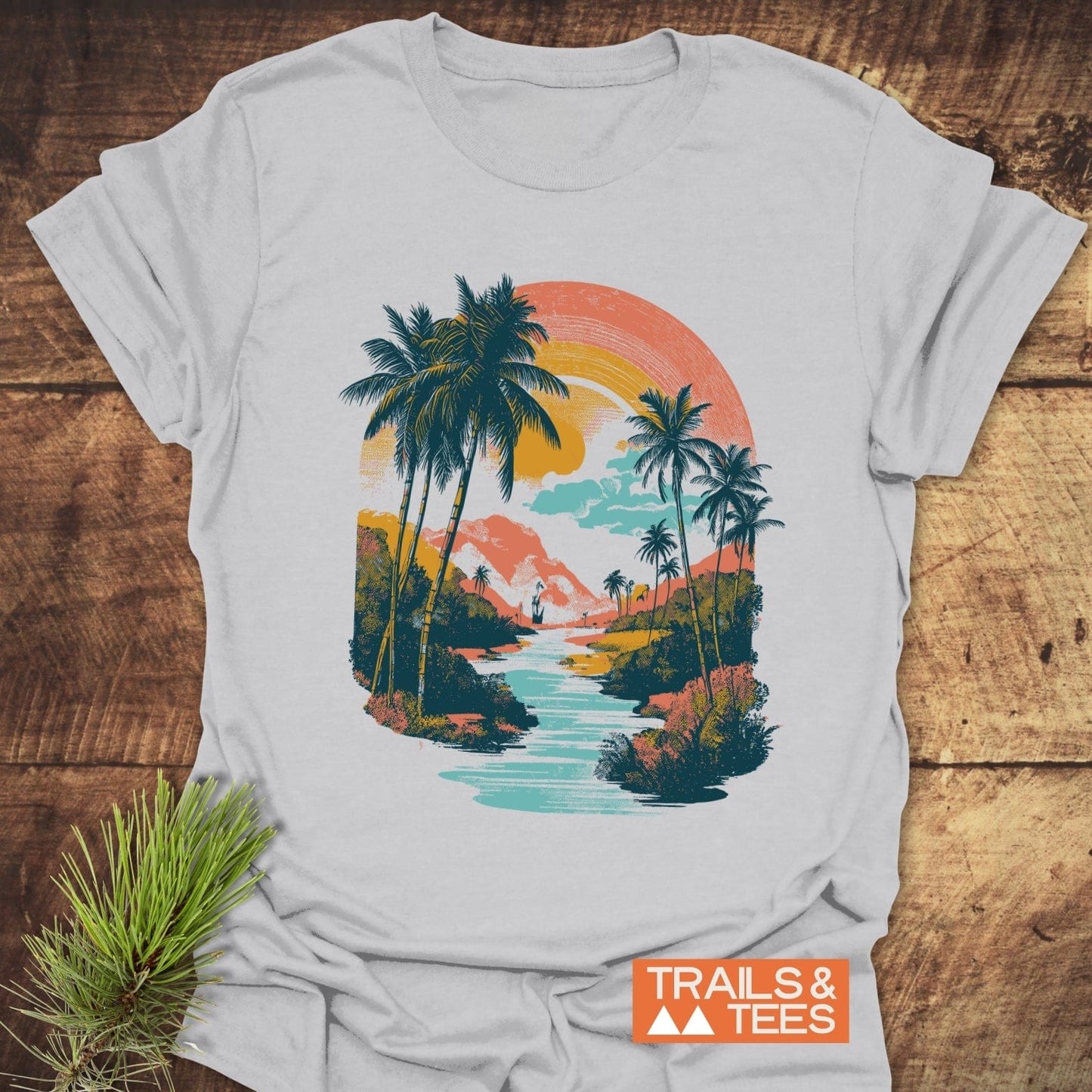 Along The River T-Shirt featuring a pastel tropical sunset scene with palm trees, crafted from soft cotton for everyday adventures. Includes TRAILS & TEES logo.