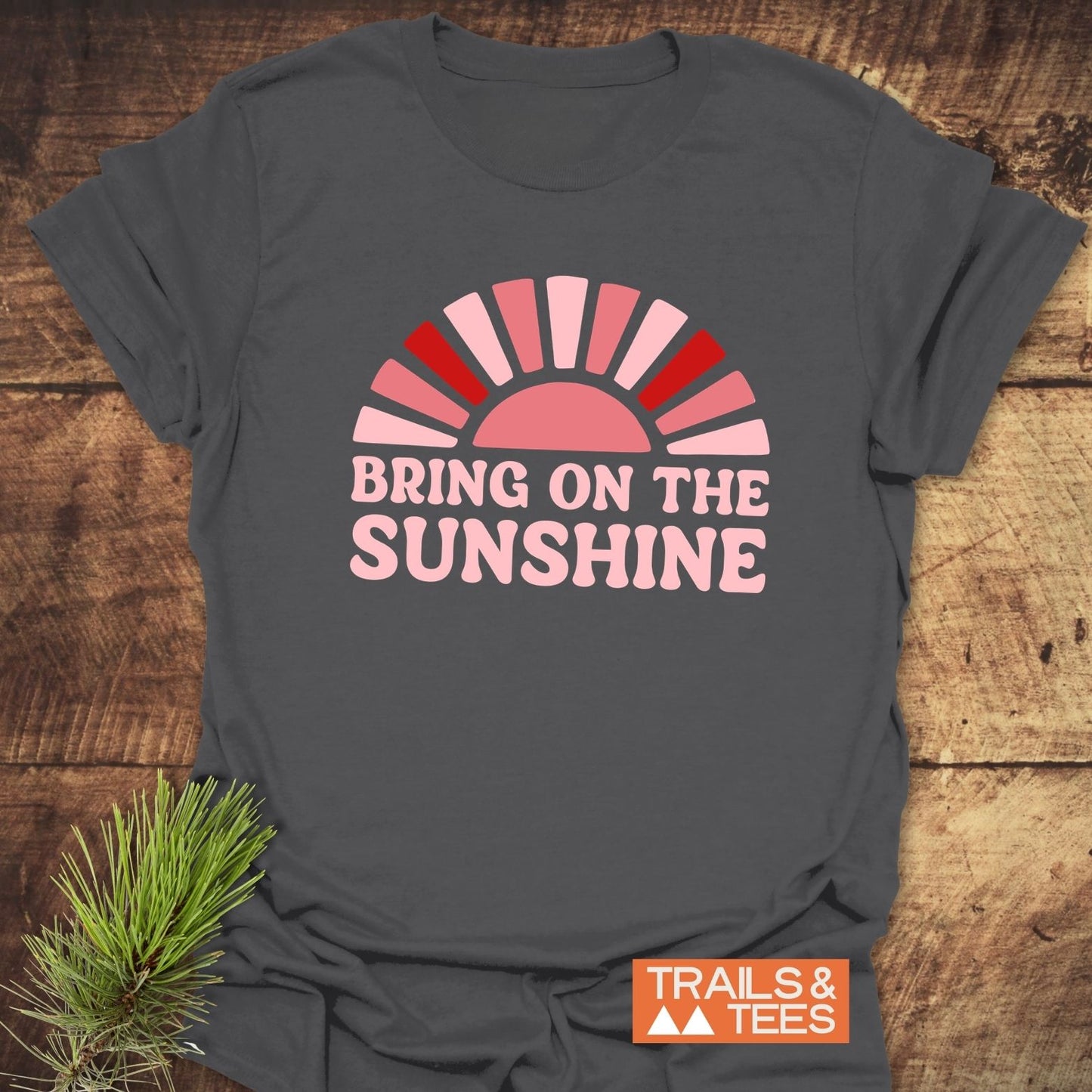Bring On The Sunshine T-Shirt featuring a pink and red half-sun graphic, captured flat-lay on wood, showcasing its soft, lightweight cotton fabric and durable print.