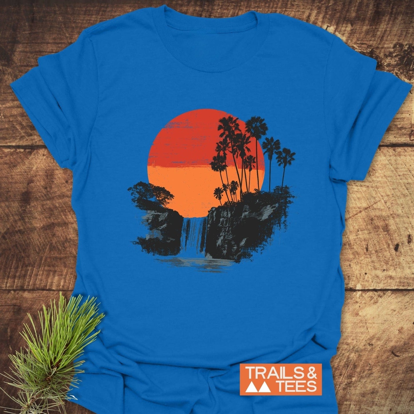 Sunset Waterfalls T-Shirt shows a tropical scene with cliffs, waterfall, and trees, featuring TRAILS & TEES logo; crafted for comfort and adventure.