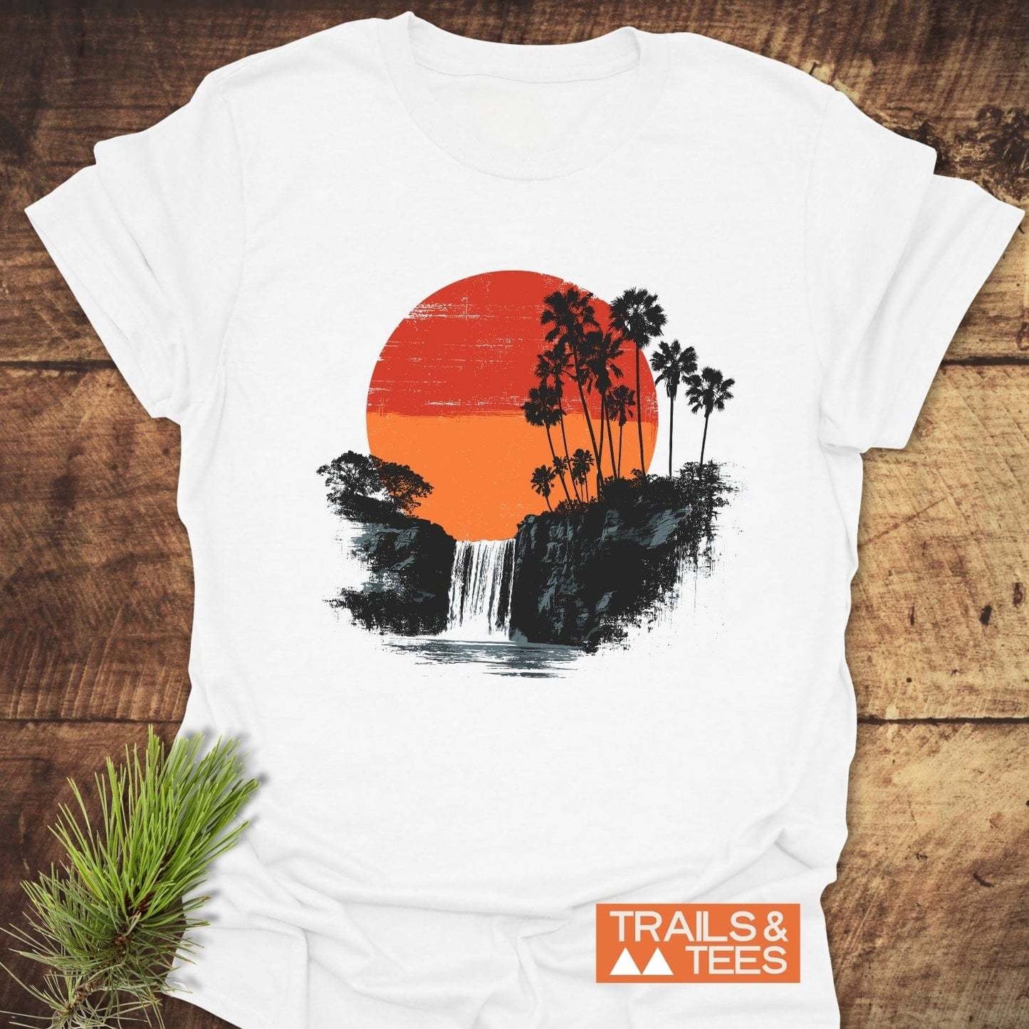 Sunset Waterfalls T-Shirt with a red-orange sun, palm trees, and waterfall design on soft cotton, laid out on wood with TRAILS & TEES logo.