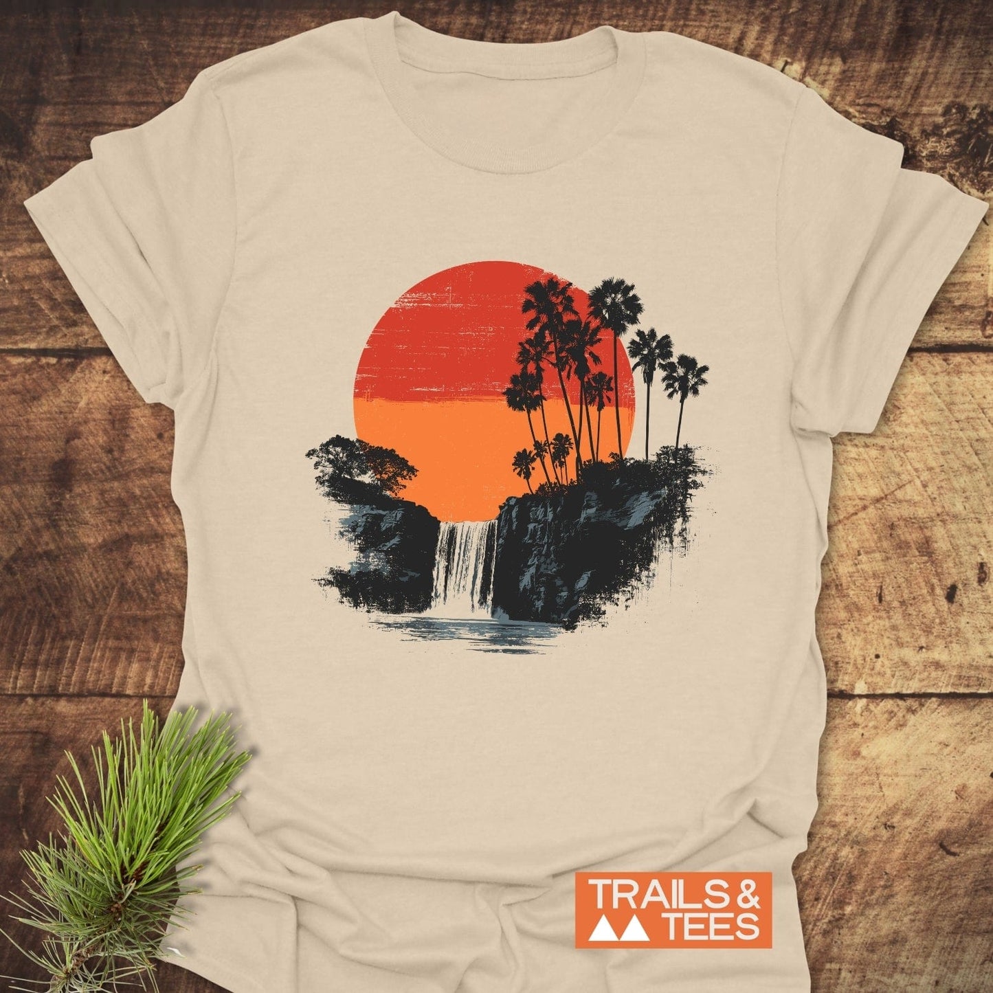 Sunset Waterfalls T-Shirt featuring a retro outdoor scene with a two-tone orange sun, cliff-edge waterfall, and black palm trees on light-beige cotton.