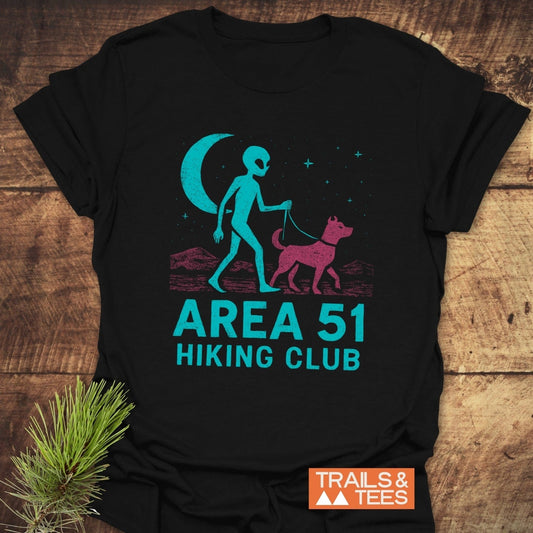 Area 51 Hiking Club T-Shirt featuring an alien walking a dog graphic, laid on wood. Crafted from soft cotton for outdoor adventures.