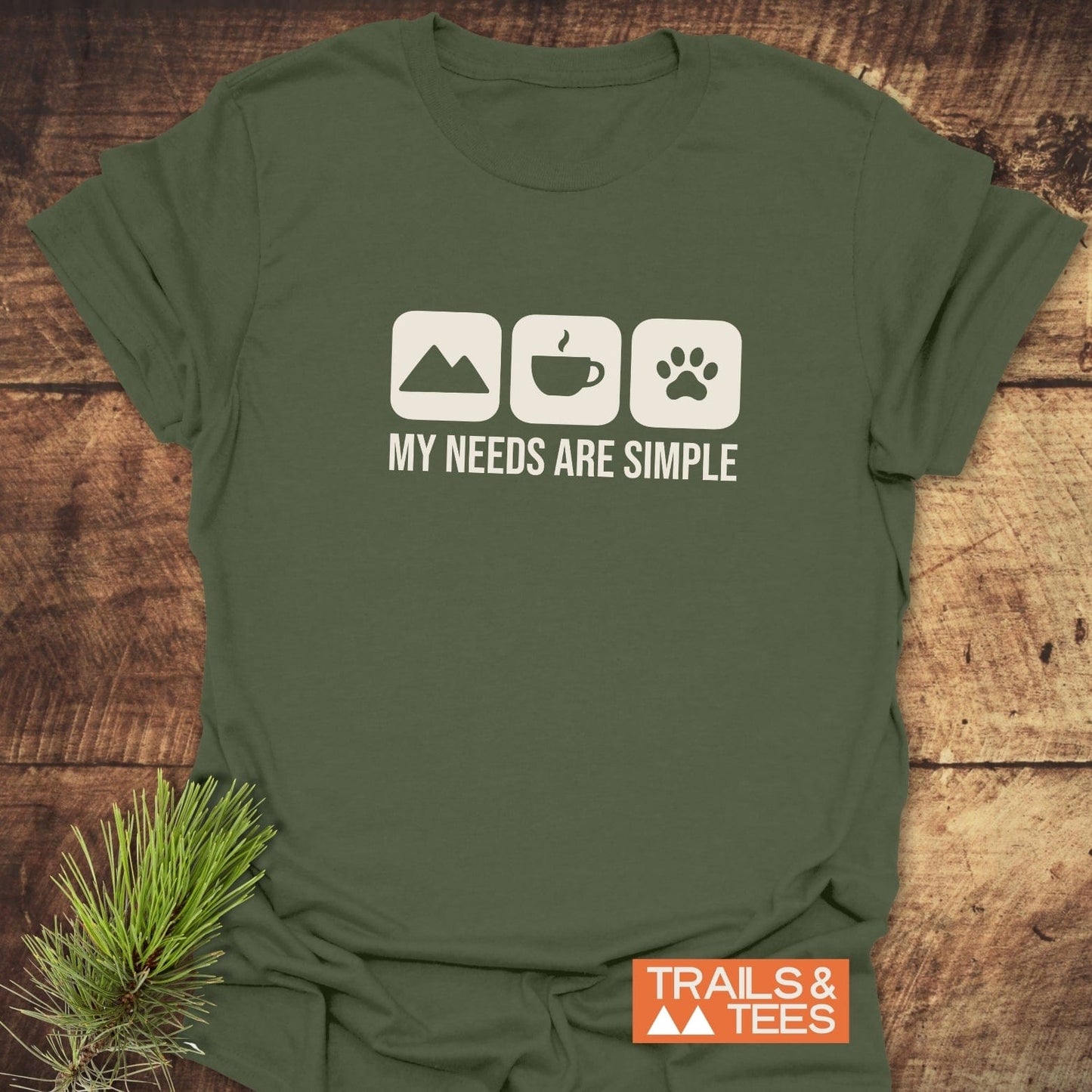 My Needs Are Simple T-Shirt with mountain, coffee, and paw icons; laid flat with a rustic touch. Soft, durable, adventure-ready cotton.