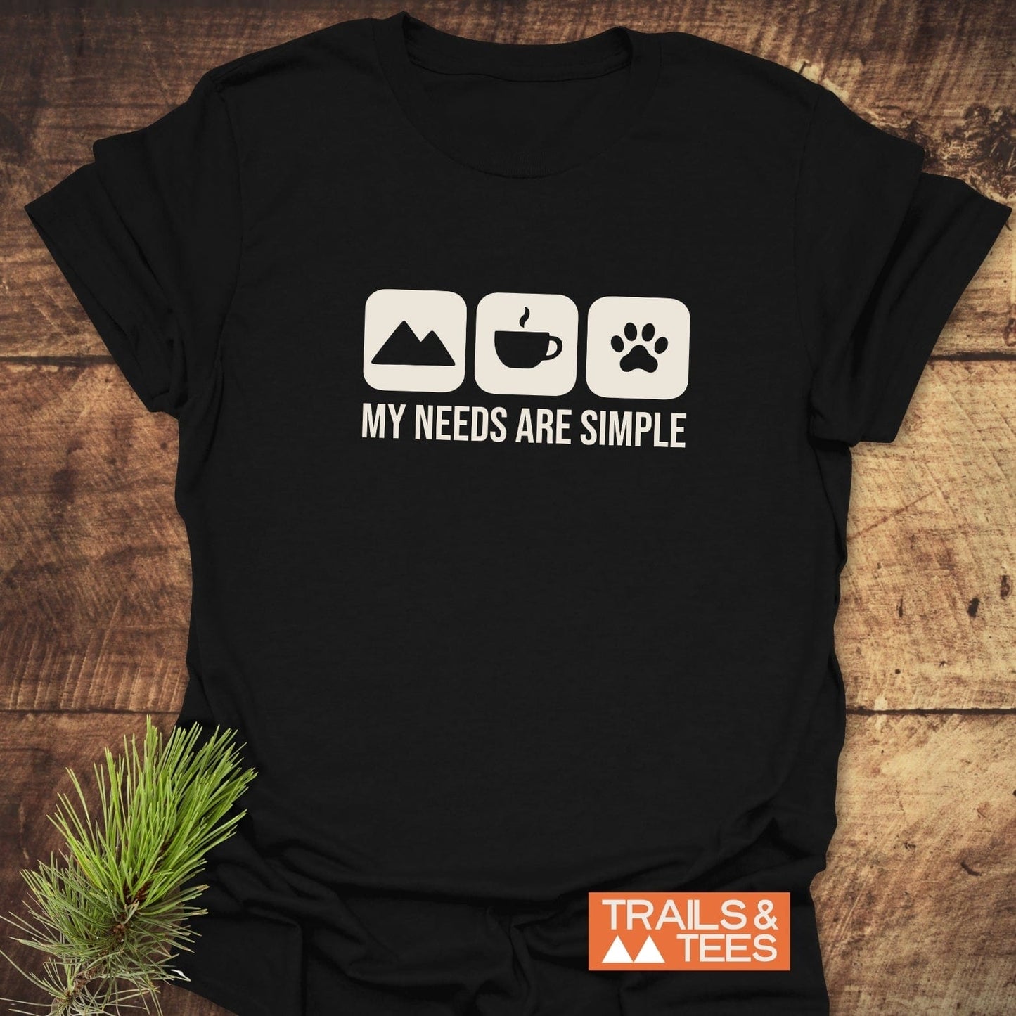 My Needs Are Simple T-Shirt with mountain, coffee, and paw icons, slogan on chest; soft cotton, durable prints, pre-shrunk, logo TRAILS & TEES visible.