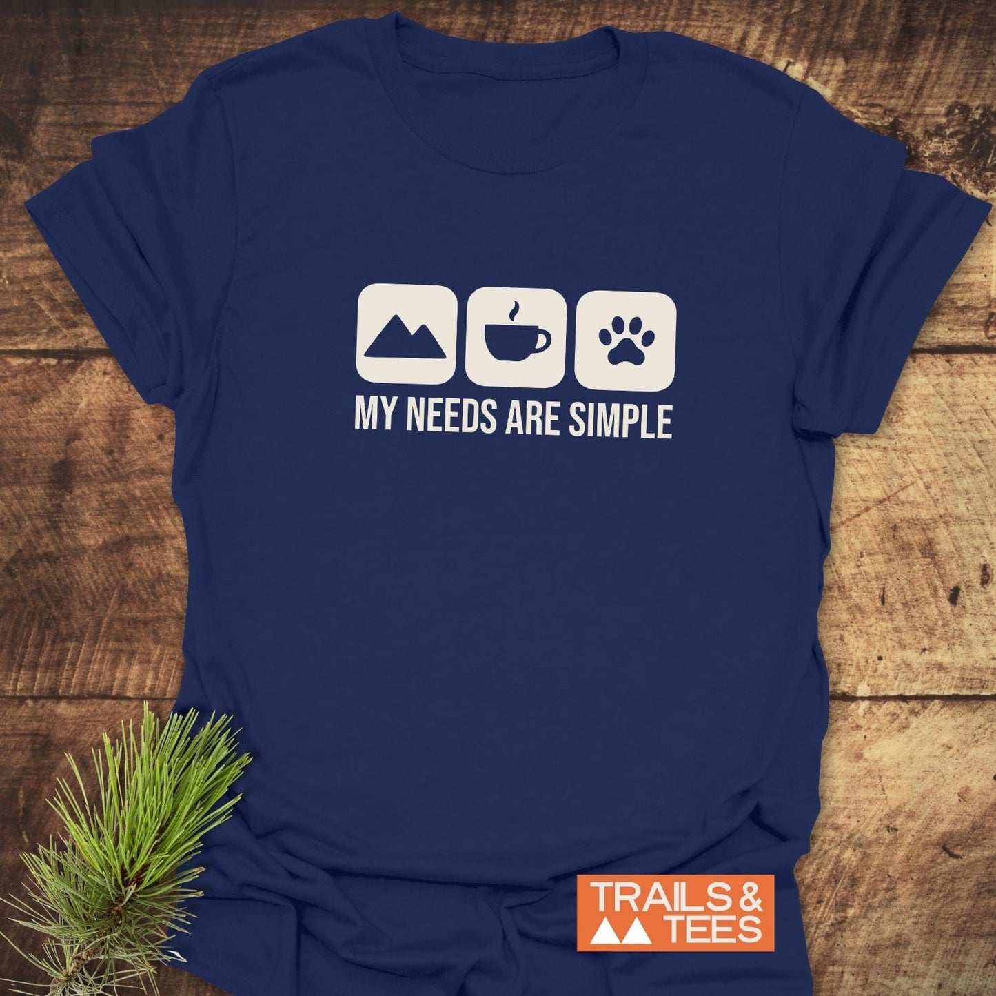 Navy-blue My Needs Are Simple T-shirt with mountain, coffee, and paw print icons, featuring TRAILS & TEES logo and styled with a pine branch.