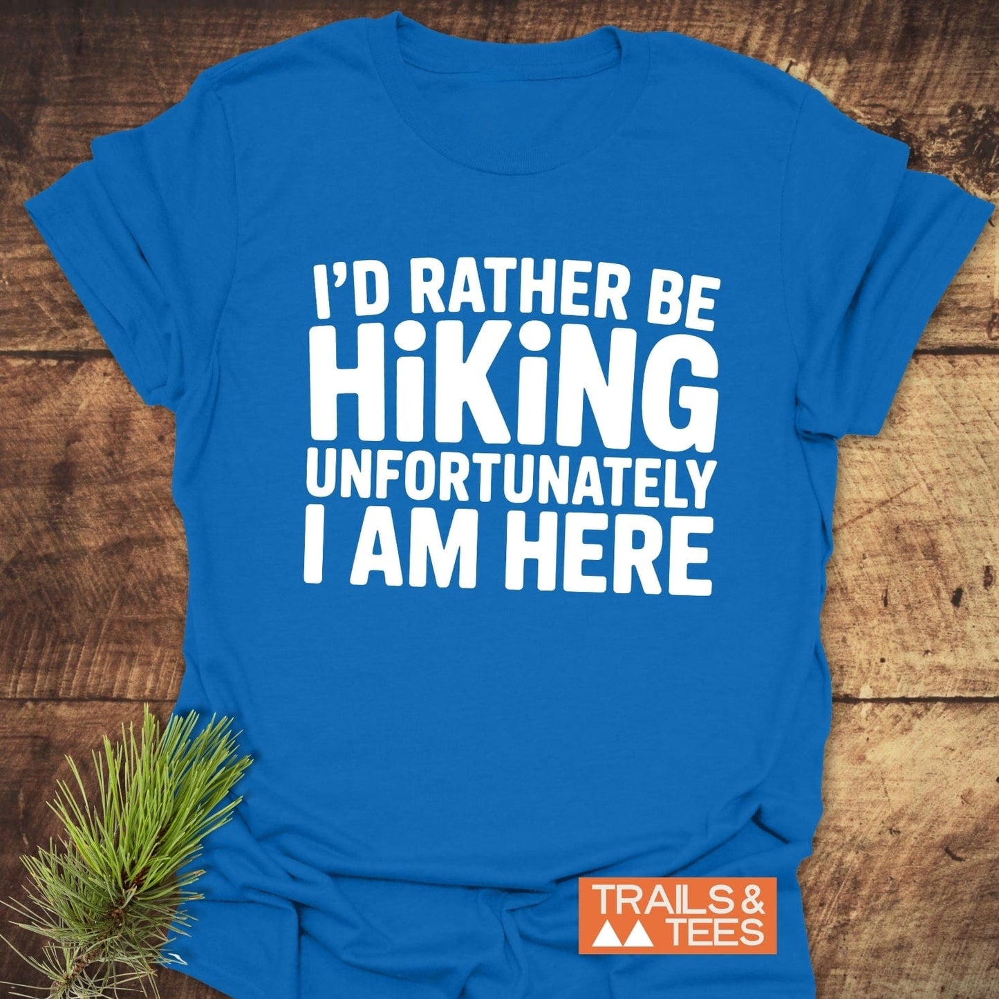 I'd Rather Be Hiking... T-Shirt, blue, with white text and an orange Trails & Tees logo, laid on wood, accented by pine needles.