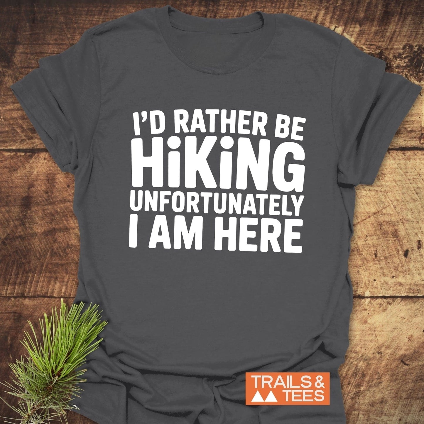 Dark-gray I'd Rather Be Hiking... T-shirt on wooden surface, featuring white lettering and TRAILS & TEES logo, highlighting comfort and durability for outdoor enthusiasts.