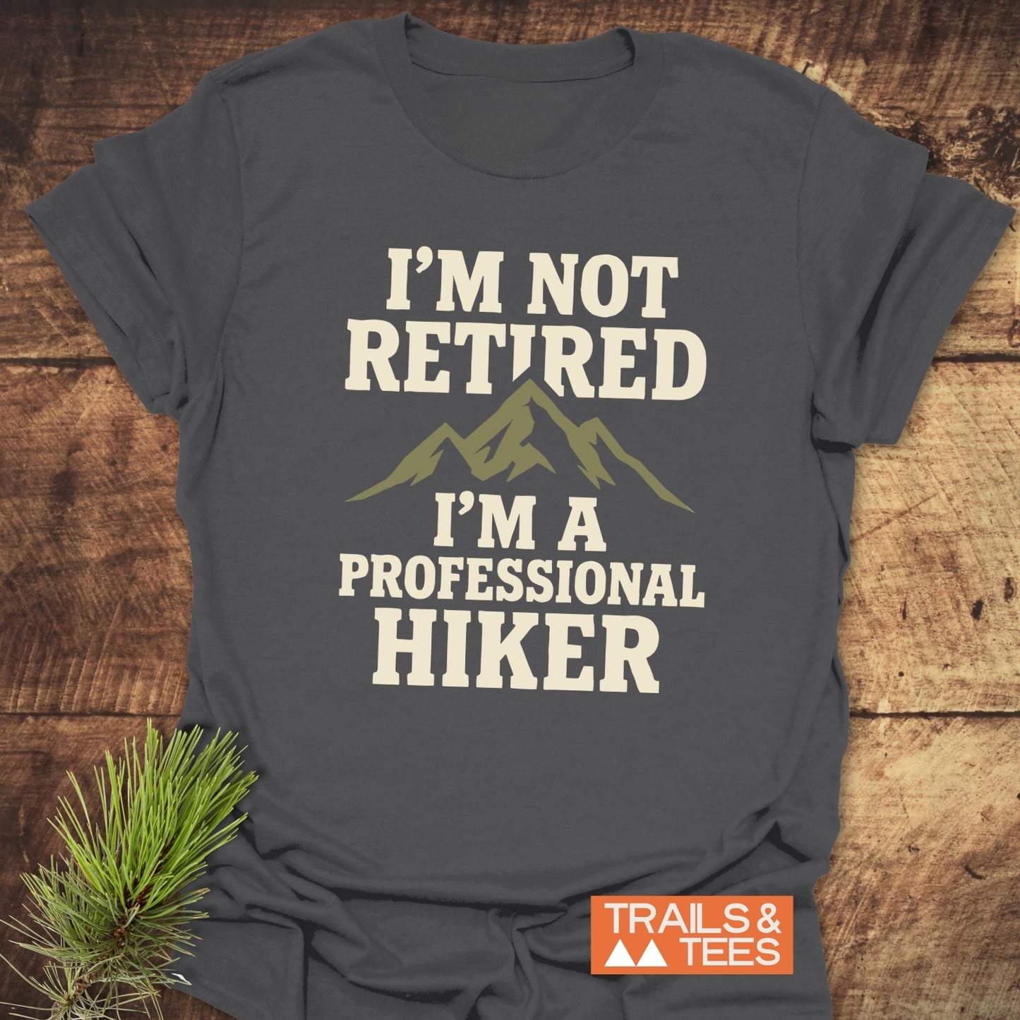 I'm Not Retired T-Shirt on wooden surface, featuring cream slogan with mountain silhouette. Designed for comfort and adventure, with durable print and pre-shrunk fit.