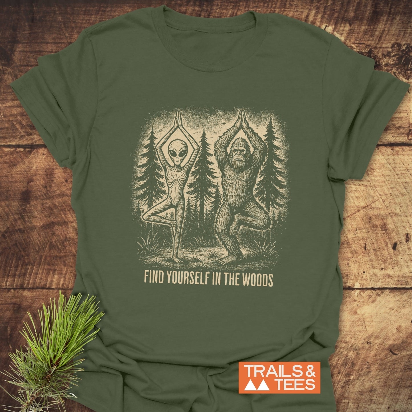 Find Yourself In The Woods T-Shirt featuring an alien and Bigfoot in tree-pose yoga stance, with slogan and Trails & Tees logo, on soft cotton.