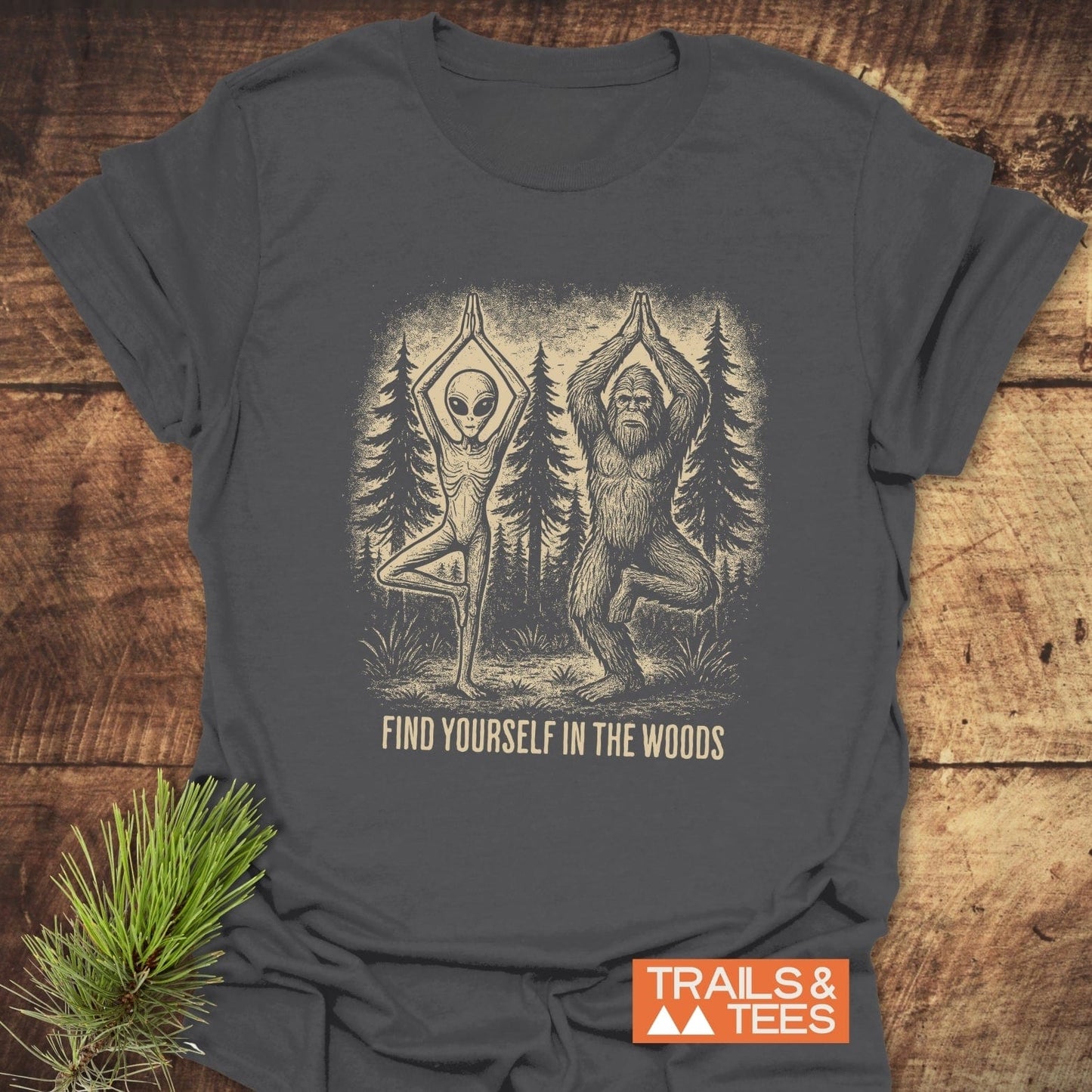 Find Yourself In The Woods T-Shirt: Flat-lay of charcoal tee featuring alien and Bigfoot in yoga pose, captioned for outdoor enthusiasts. Made from soft, durable, ring-spun cotton.