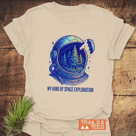 My Kind Of Space Exploration T-Shirt featuring a neon-blue astronaut helmet with pine trees inside, laid flat with a TRAILS & TEES logo and pine needles.