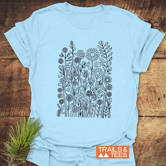 Wildflowers T-Shirt featuring a black floral design on a light-blue fabric, laid on a wood surface with a Trails & Tees logo and pine sprig accent.