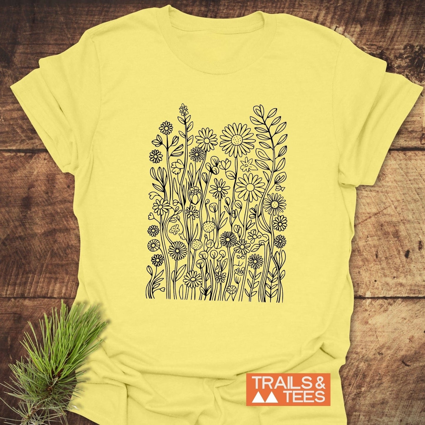 Wildflowers T-Shirt featuring hand-drawn flower illustration, laid on wood with a pine branch. Includes TRAILS & TEES logo, offers comfort and durability.