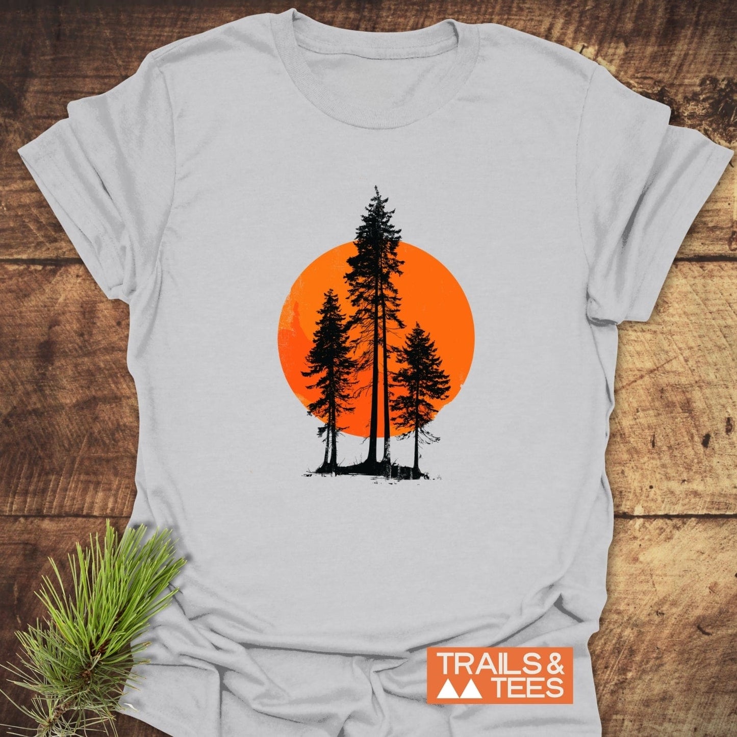 Sunset Trees T-Shirt featuring black silhouette evergreens against an orange circle, laid on wood with pine sprig and TRAILS & TEES logo nearby.
