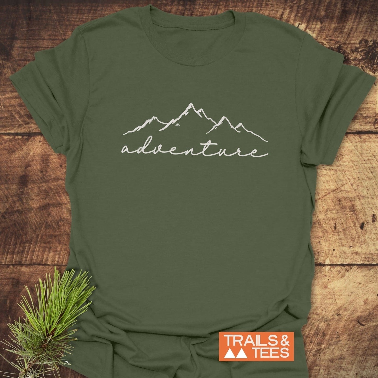 Adventure T-Shirt with mountain design in white, laid on wood. Includes TRAILS & TEES logo and pine branch accent. Soft, pre-shrunk cotton for outdoor enthusiasts.