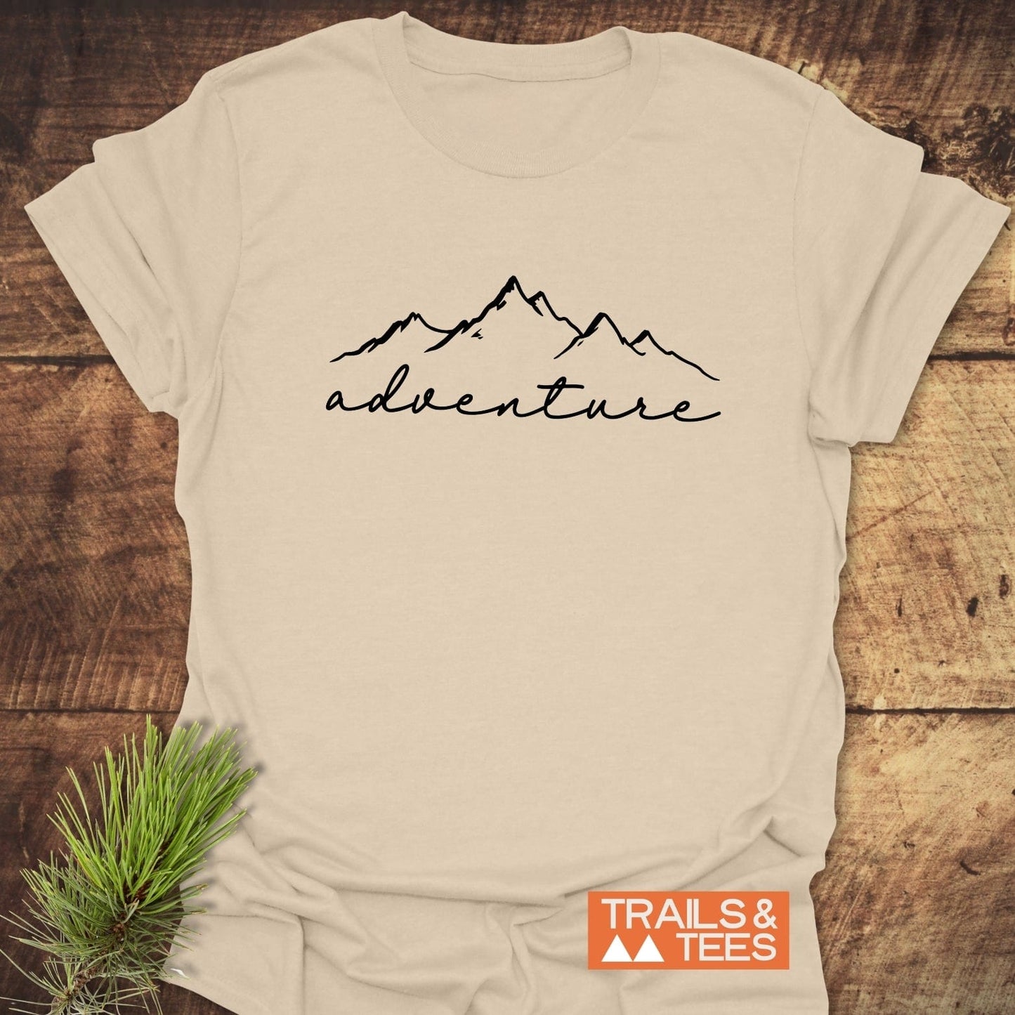 Adventure T-Shirt featuring mountain outline and adventure script on light-beige fabric, laid on wood with pine branch. Durable, comfortable, pre-shrunk cotton.