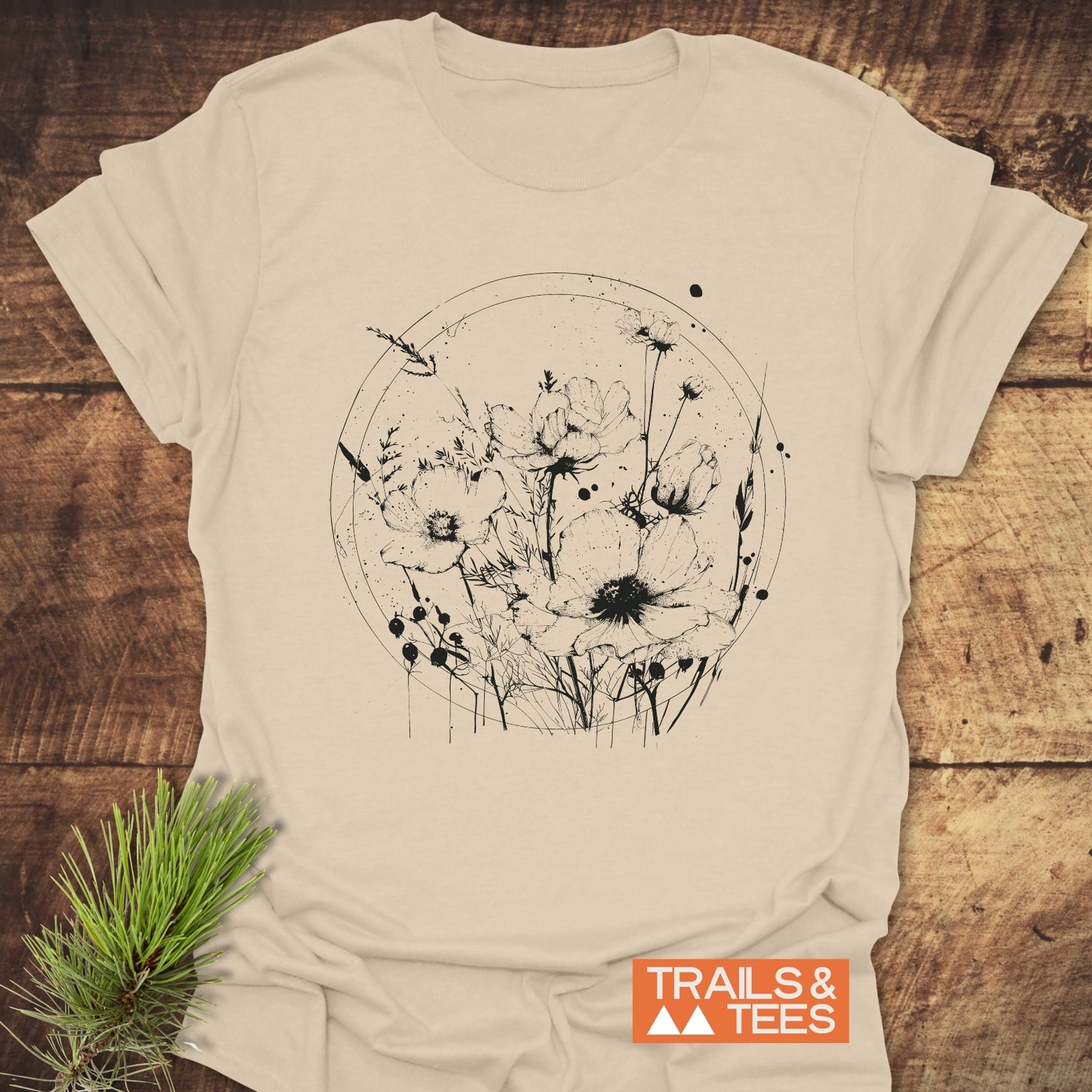 Wildflower Circle T-Shirt on wooden surface, featuring a black hand-drawn floral circle print. Includes TRAILS & TEES logo and pine sprig for styling.