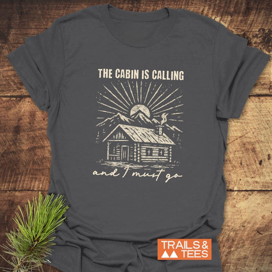 The Cabin Is Calling T-Shirt with a log cabin design, mountain backdrop, and sunbeams, crafted from soft ring-spun cotton for outdoor enthusiasts.