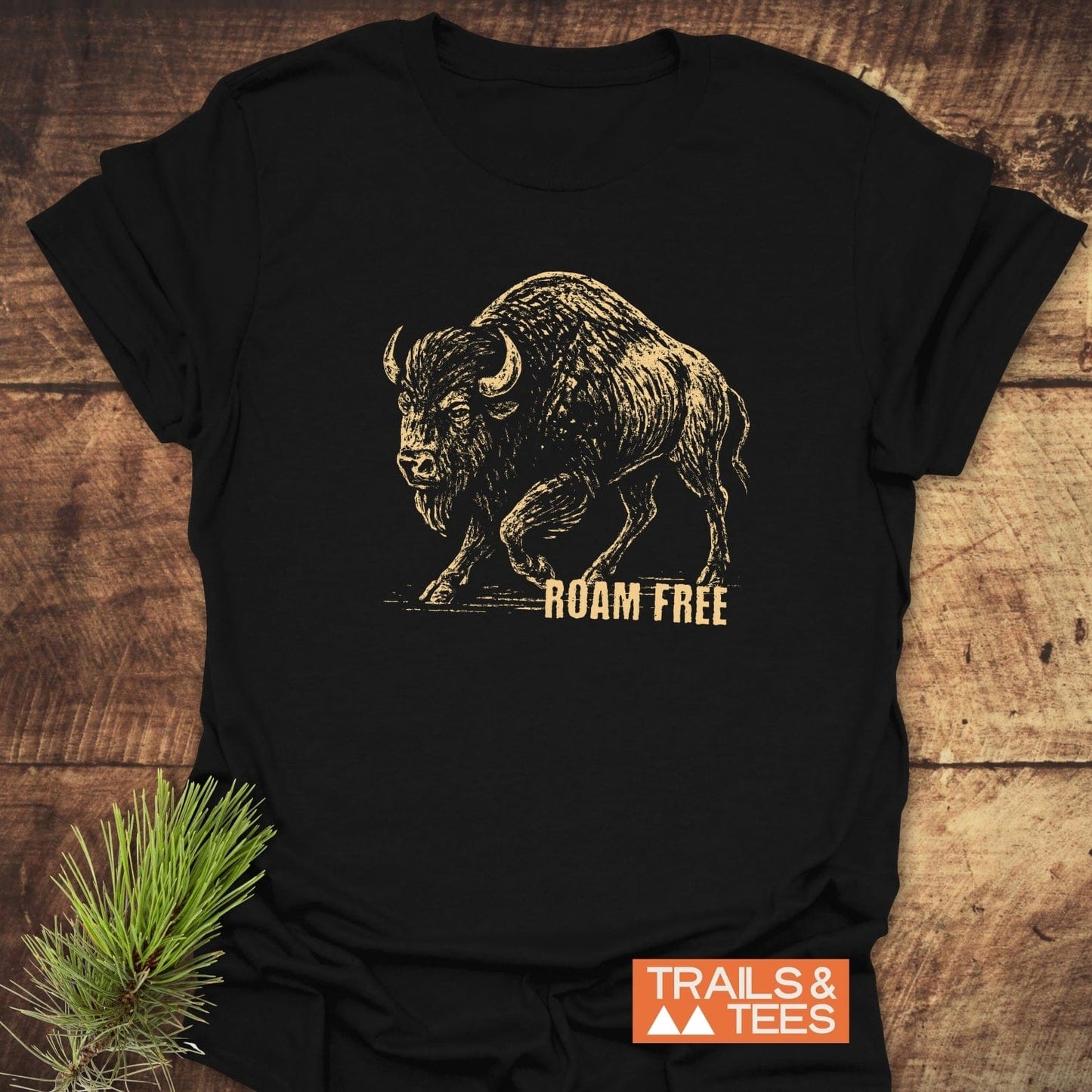 Roaming Bison T-Shirt featuring a sketch-style bison graphic and ROAM FREE slogan, styled on wood with pine, showcasing Trails & Tees logo.