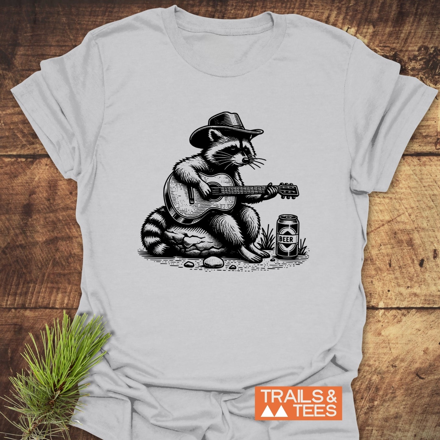 Guitar Playing Raccoon T-Shirt featuring a raccoon in a cowboy hat playing guitar, with a TRAILS & TEES logo, on soft, durable cotton.