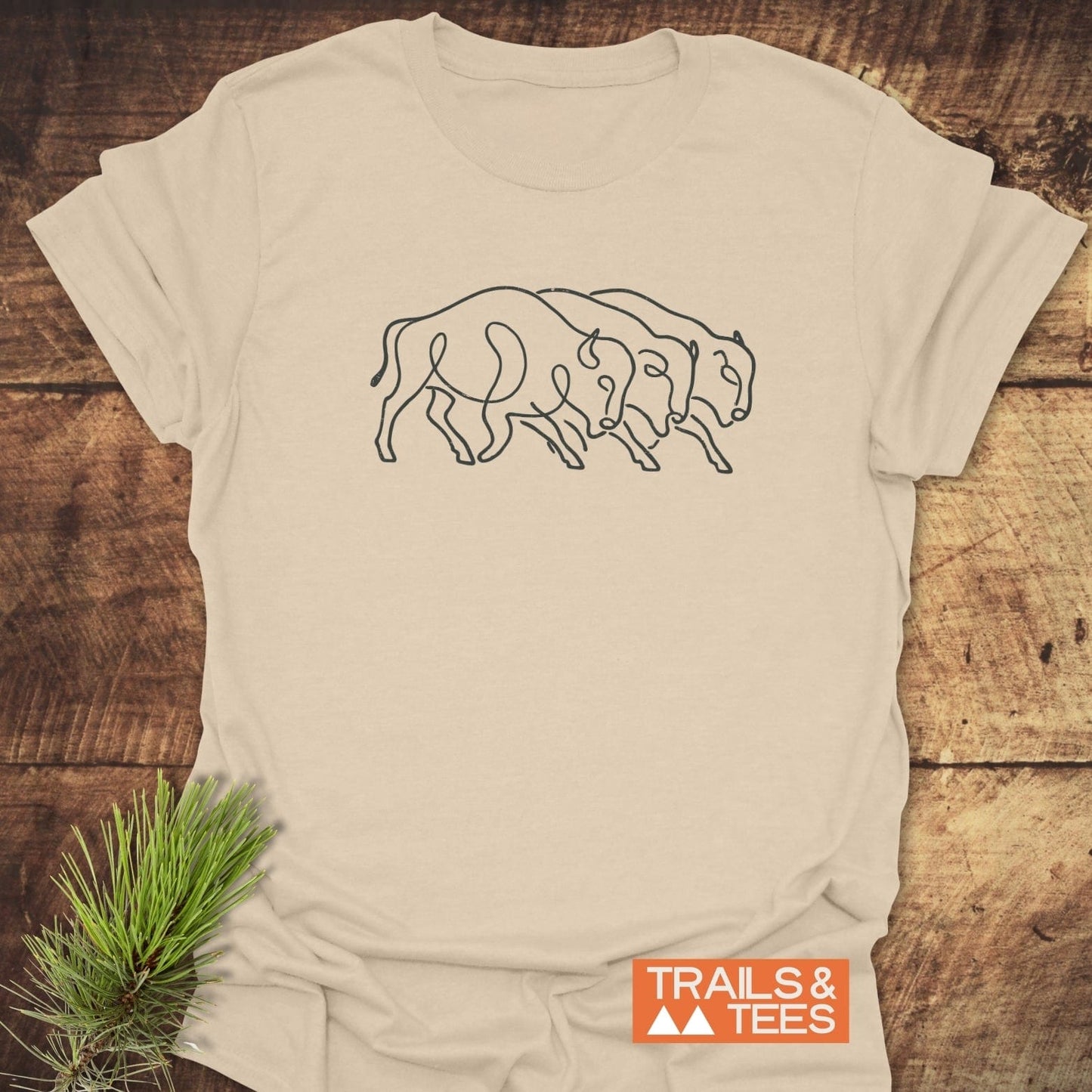 Minimalist Bison T-Shirt on rustic wood, featuring three bison illustration. Includes “Trails & Tees” logo. Lightweight, ring-spun cotton for comfort and durability.