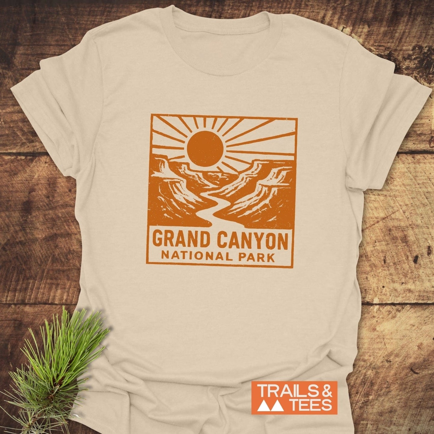 Grand Canyon NP T-Shirt on wood surface, featuring an orange sun and canyon graphic. Includes TRAILS & TEES logo, pine sprig accentuates outdoor theme.