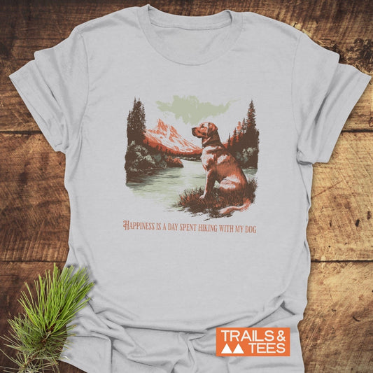 Happiness Is Hiking With Dogs T-Shirt featuring a mountain lake scene with a dog, evergreen trees, and peaks. Made from soft, durable cotton by TRAILS & TEES.