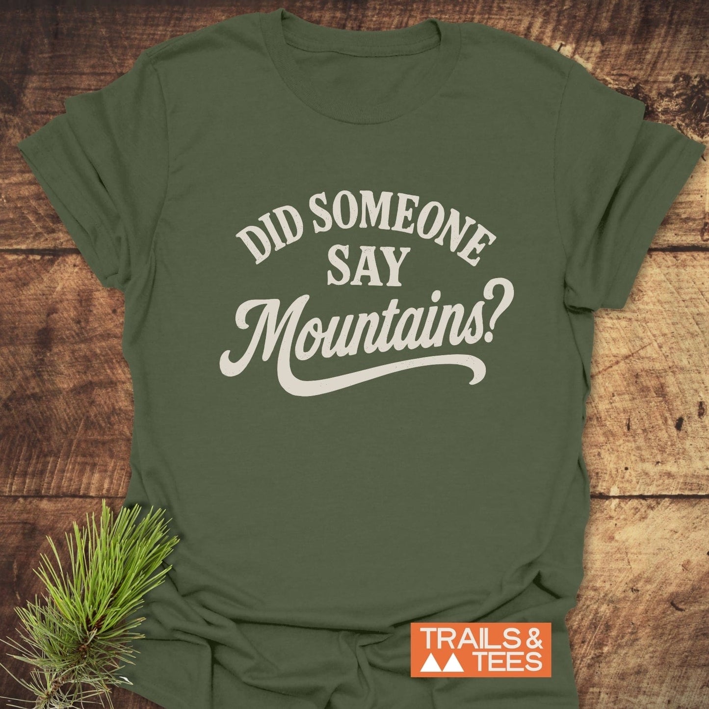 Forest-green 'Did Someone Say Mountains' T-Shirt on wood, featuring off-white text, orange 'TRAILS & TEES' logo, and pine sprig, embodying outdoor adventure spirit.