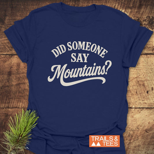Navy-blue Did Someone Say Mountains? T-shirt with white lettering, orange TRAILS & TEES logo, styled with pine sprig on wooden surface.