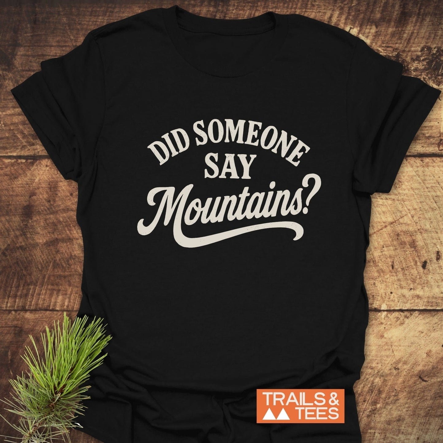 Black T-shirt with “DID SOMEONE SAY Mountains?” and “TRAILS & TEES” logo, laid on wood with pine branch, embodies outdoor adventure spirit.