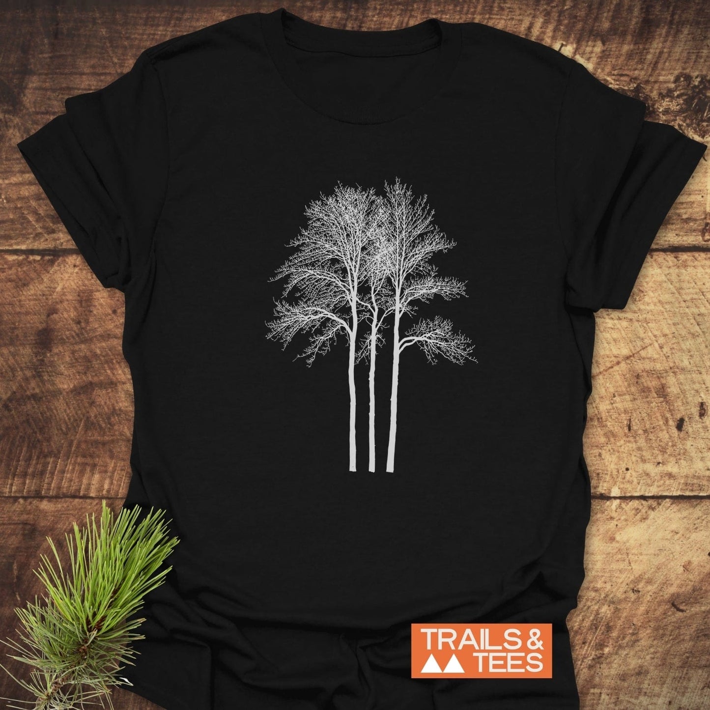 Tree Silhouettes T-Shirt on wood surface, featuring white tree graphic. Includes “TRAILS & TEES” logo. Made from soft cotton, durable print, pre-shrunk for reliable fit.