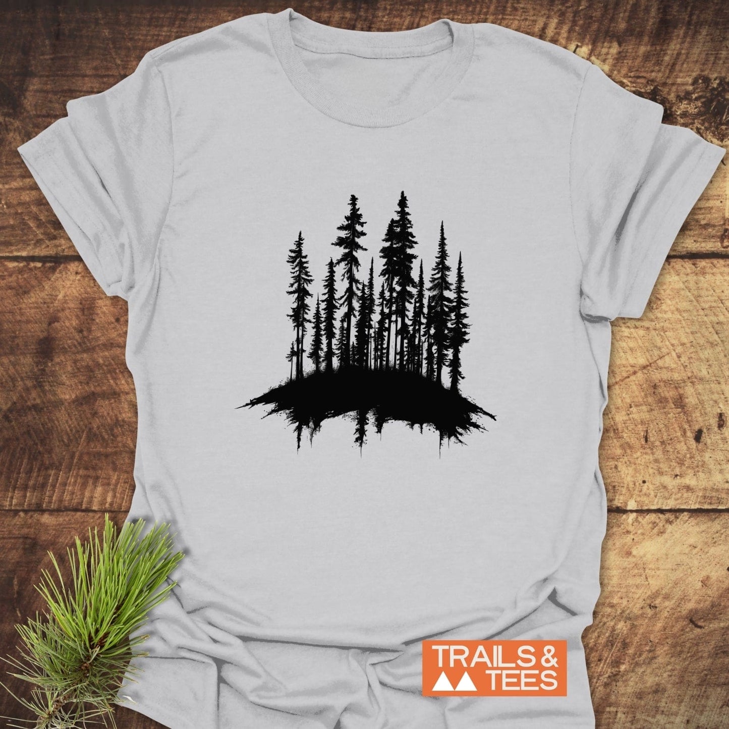 Woods Silhouette T-Shirt featuring a black evergreen tree cluster design on light gray fabric, displayed with a pine sprig, emphasizes outdoor adventure and comfort.