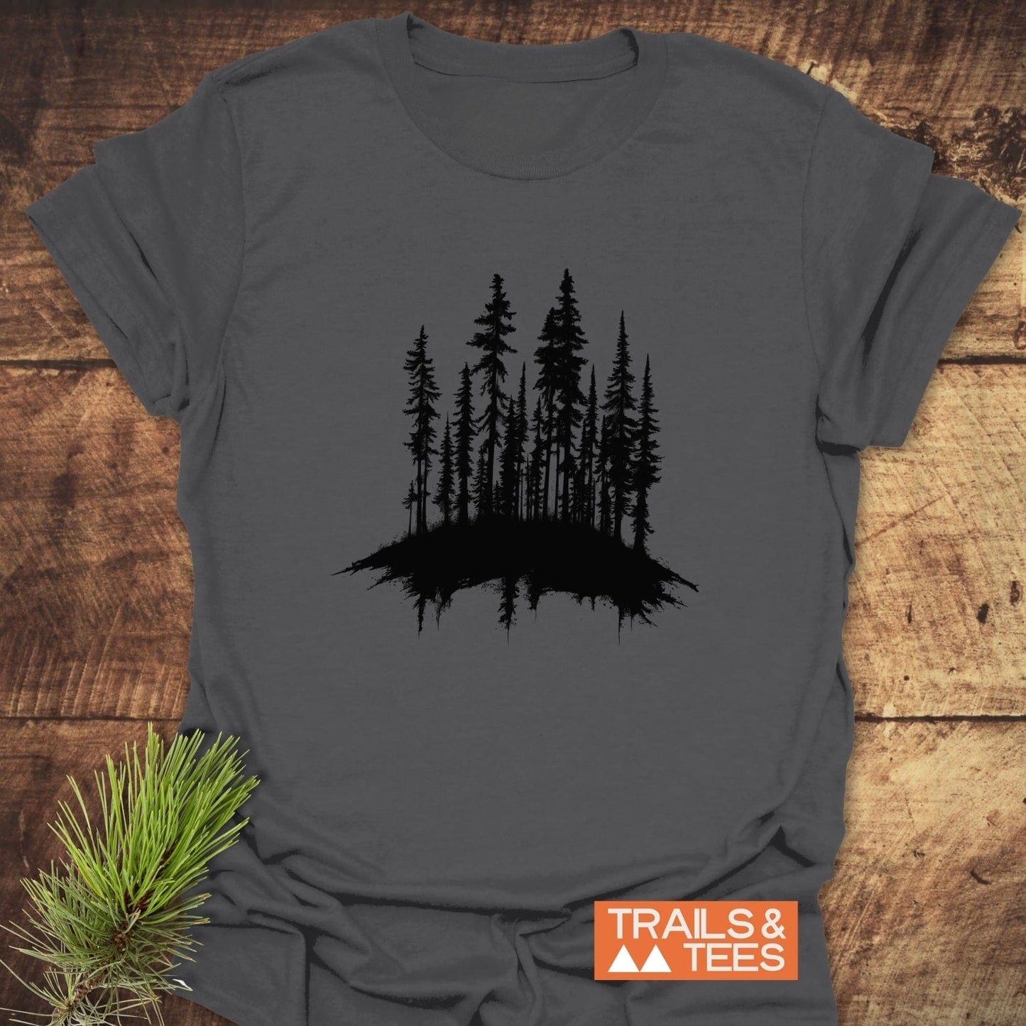Woods Silhouette T-Shirt on wooden surface, featuring black pine tree design. Includes TRAILS & TEES logo and pine branch. Soft, durable, pre-shrunk cotton for outdoor adventures.