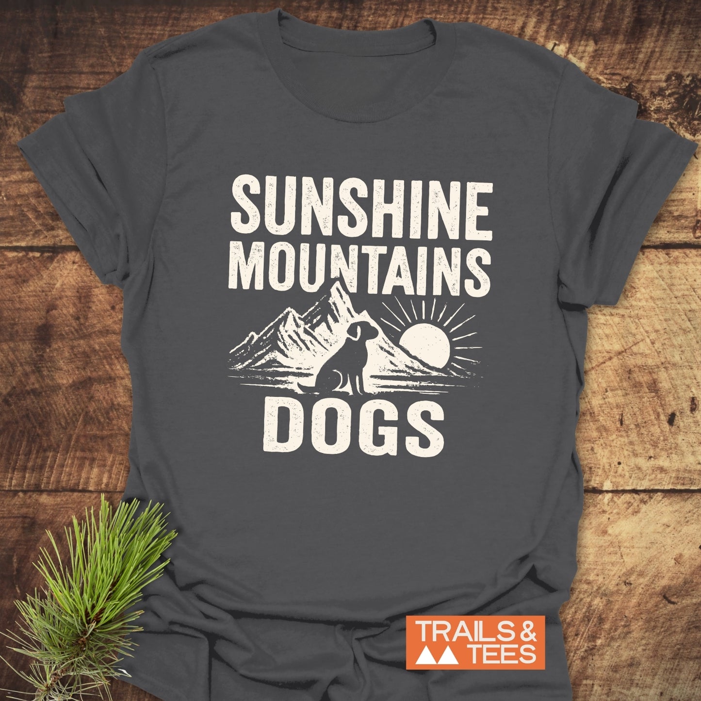Sunshine Mountains Dogs T-Shirt on wood, featuring a dog silhouette with mountains and sun graphic. Includes TRAILS & TEES badge, highlighting outdoor adventure theme.
