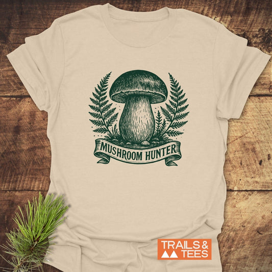 Mushroom Hunter T-Shirt with a green mushroom illustration, fern fronds, and “TRAILS & TEES” logo, laid on wood. Made from soft, durable ring-spun cotton.
