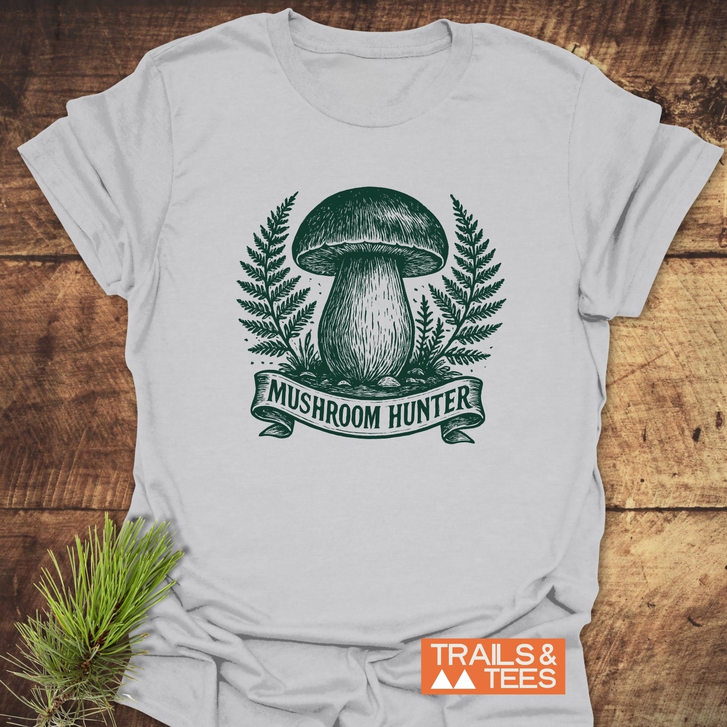 Mushroom Hunter T-Shirt on wood surface, featuring green mushroom and fern design, with a pine sprig nearby. Perfect for outdoor enthusiasts.