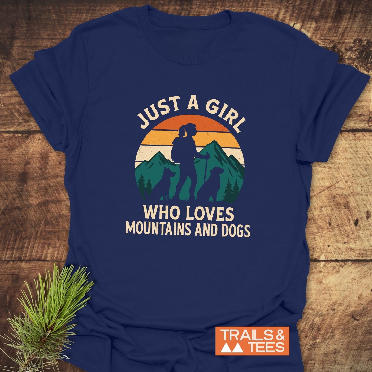 Mountains And Dogs T-Shirt featuring a hiker and dogs graphic, slogan, and TRAILS & TEES logo, laid on a wooden surface.