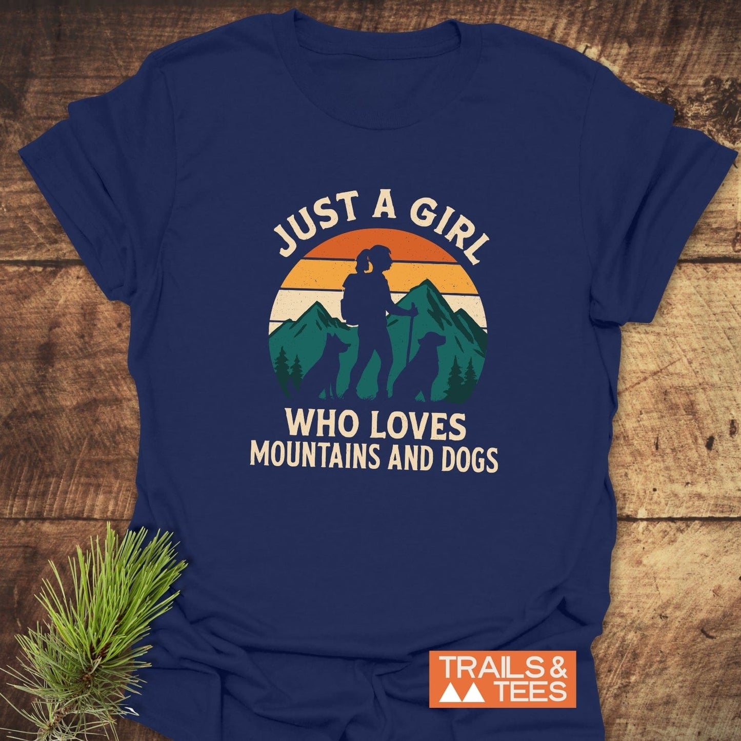 Mountains And Dogs T-Shirt featuring a hiker and dogs graphic, slogan, and TRAILS & TEES logo, laid on a wooden surface.