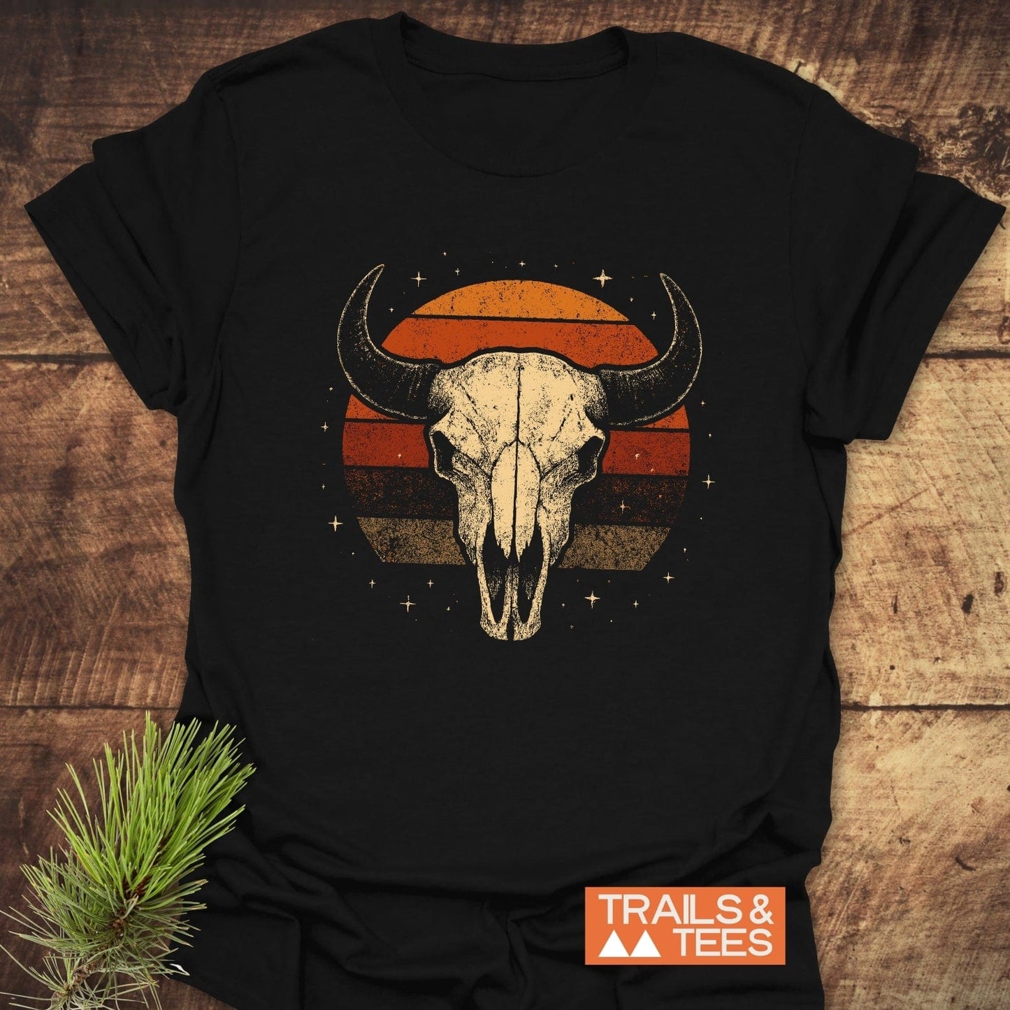 Retro Buffalo Skull T-Shirt on wood with vintage skull and sunset graphic. Soft, lightweight cotton, pre-shrunk, durable print. Label: TRAILS & TEES. Pine sprig beside.