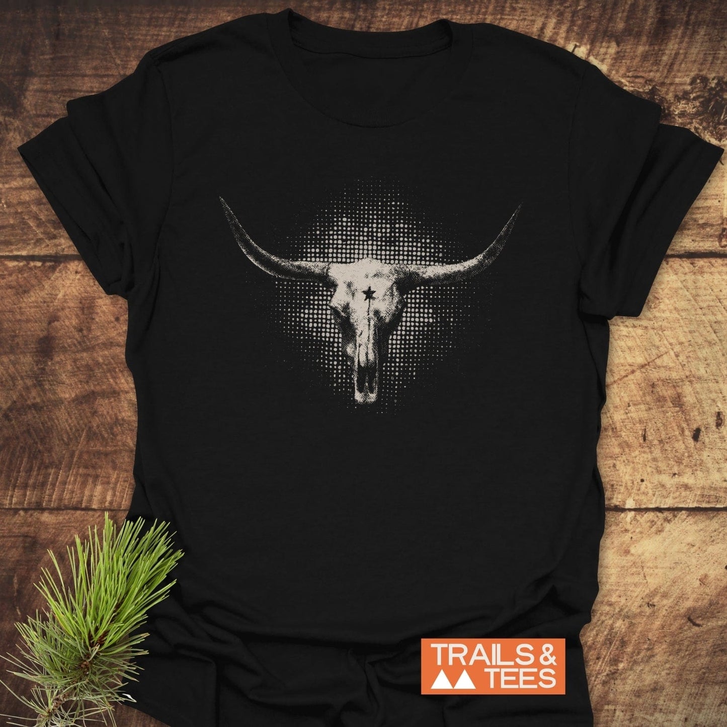 Buffalo Skull Texas T-Shirt featuring a white longhorn skull graphic on a black tee, laid on wood with a pine twig and TRAILS & TEES logo.